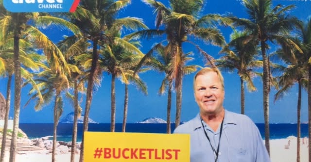 Kirk Webster beside a bright #BUCKETLIST sign among palm trees on a beach